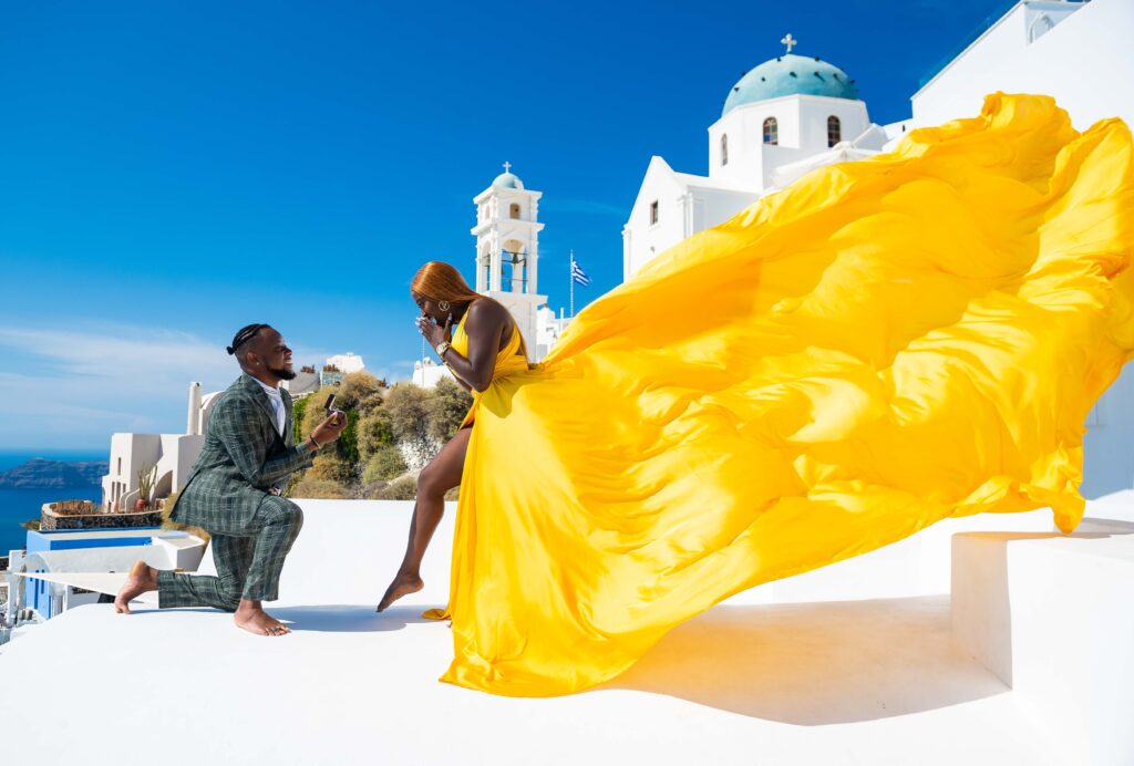 Man proposing on one knee to a woman in a yellow flying dress beside a blue-domed church