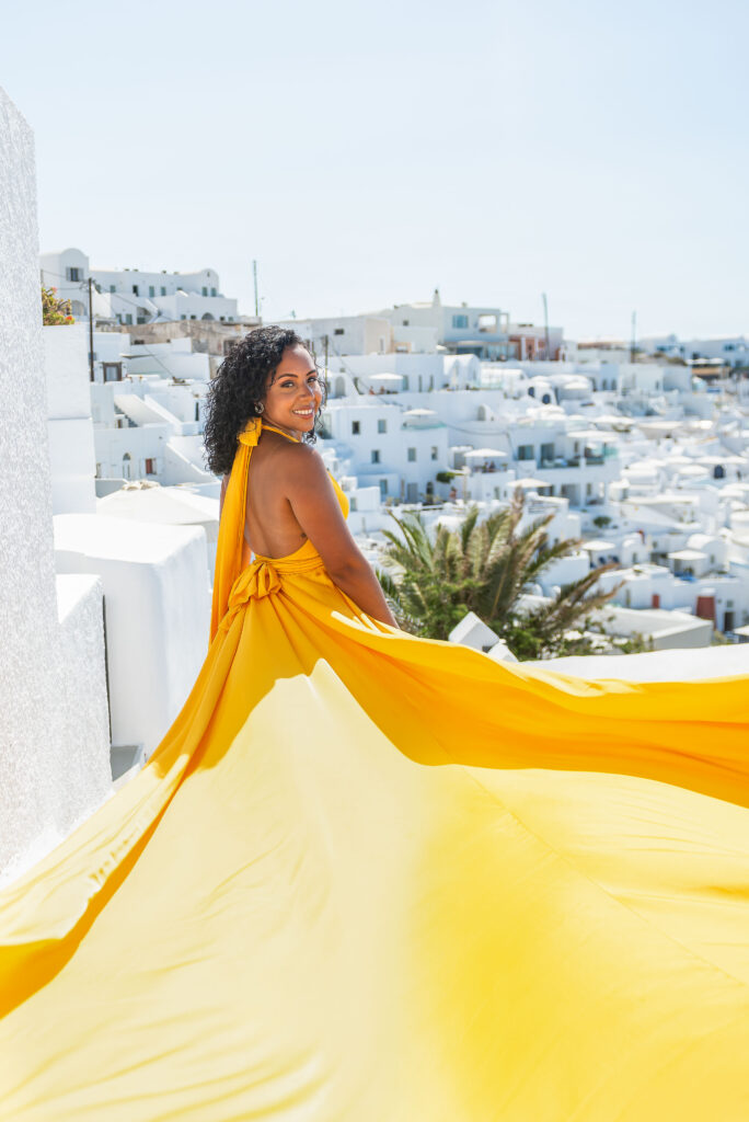 Smiling woman looking back in a yellow flying dress above Santorini’s whitewashed rooftops