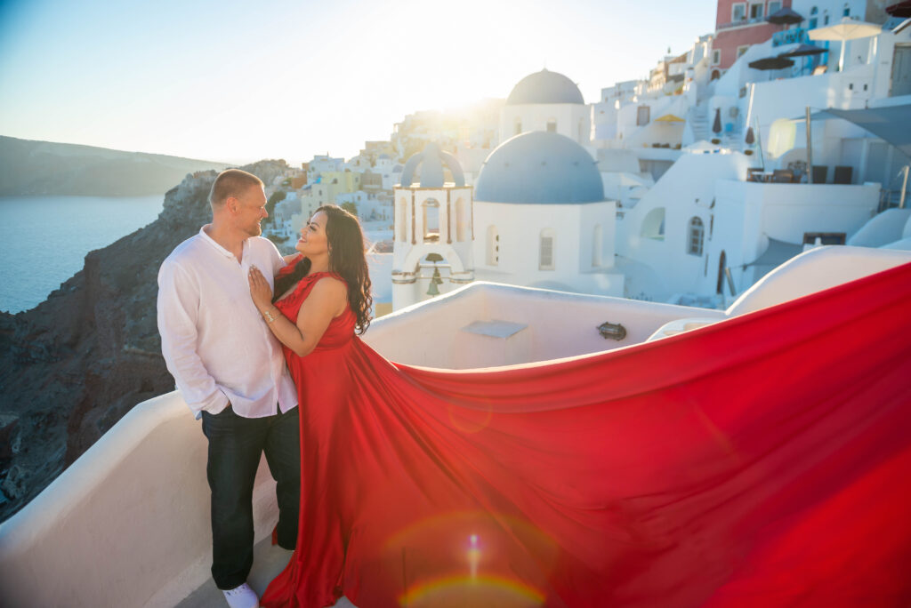 A couple standing together with a wonderful Santorini sunset. They're looking at each other and smiling. In the back we see white roofs and blue dome church.