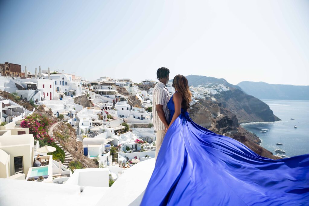 A couple poses for a photoshoot against the stunning backdrop of the white houses in Oia, with the lady wearing a flowing blue dress that gives the image a sense of movement and grace. The romantic setting and the couple's affectionate poses capture the essence of love and beauty, creating a timeless and enchanting image.