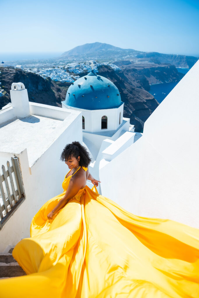 A stunning photo captures a woman dressed in yellow walking down a flight of stairs, with a top angle view of the iconic Santorini scenery in the background. The contrast between the woman's bright outfit and the white and blue tones of the surrounding buildings creates a visually appealing image that showcases the beauty of this destination