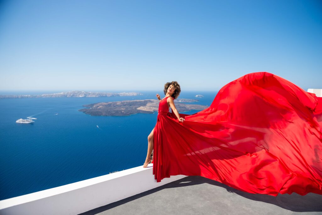 A woman stands on the edge of a scenic cliff, wearing a vibrant red flowing dress that stands out against the blue hues of the sky and sea. Behind her lies the breathtaking view of the Santorini volcano, adding to the drama and beauty of the moment