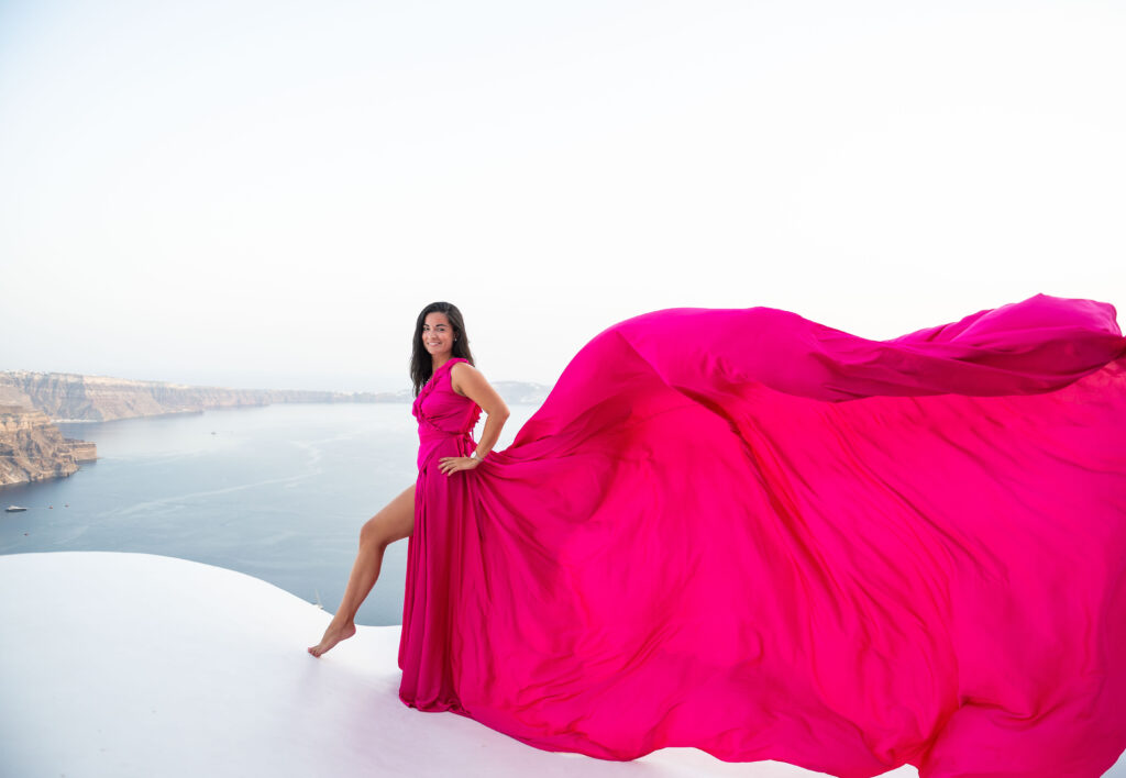 A woman strikes a pose with her hand on her wrist, donning a striking fuchsia dress with ruffled detailing. Her dress features a long trail that cascades behind her as she looks out at a stunning view of an island landscape