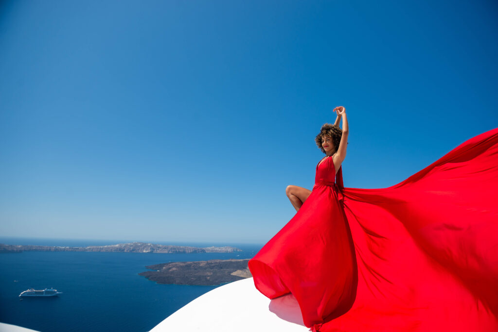 An elegant lady wearing a flowing red dress gracefully dances on a bright white rooftop with outstretched arms, exuding a sense of freedom and joy