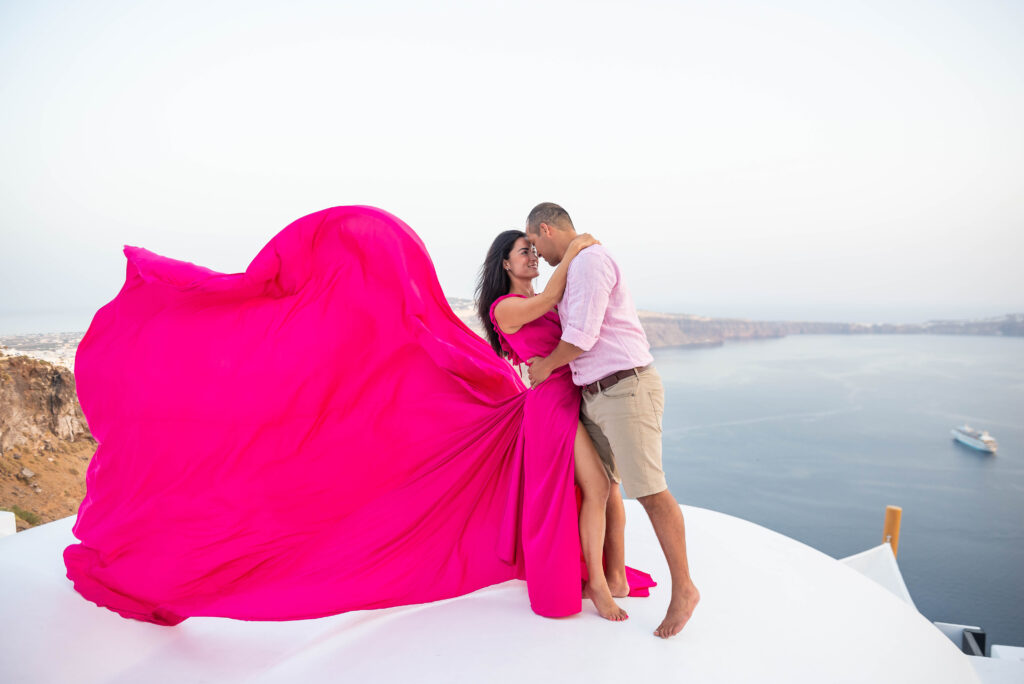 A couple standing on top of Rocco mare hotel. They are hugged and looking each other, a woman wears a pink flying dress and a man nice shorts and pink top.