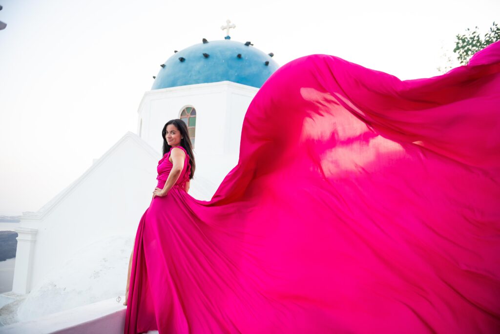In this image, a woman is wearing a beautiful fuchsia dress with ruffled details while standing close to the Church of Anastasi in Imerovigli. The church is a white, traditional Cycladic style building with a blue dome and is situated on a rocky hill overlooking the Aegean Sea. The contrast between the bright pink of the woman's dress and the white and blue of the church creates a striking image.