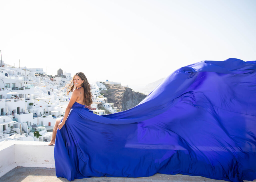 A woman in a flowing blue dress is standing on a rooftop, overlooking a breathtaking view of white houses. The houses are perched on the side of a cliff, with the sparkling blue waters of the Aegean Sea stretching out behind them. The woman's dress appears to be caught in a gentle breeze, giving the impression that she might take flight at any moment.