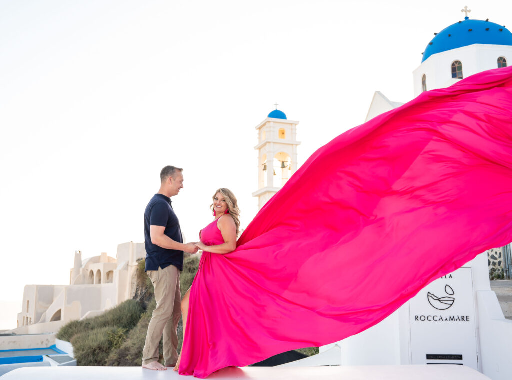 A scenic view of a couple standing on a rooftop terrace in Santorini, with a lady in a stunning fuchsia dress adding a pop of color to the beautiful surroundings.