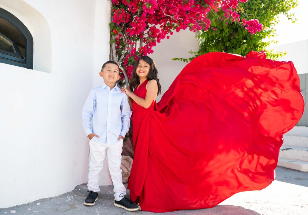 A girl and a boy are standing outdoors, with the girl wearing a colorful flying dress and the boy dressed in casual clothes. They are standing near a flowering bougainvillea plant, and the girl is holding a bouquet of flowers. Both children have joyful expressions on their faces, and the bright colors of the flowers and dress add to the playful and cheerful atmosphere of the image.