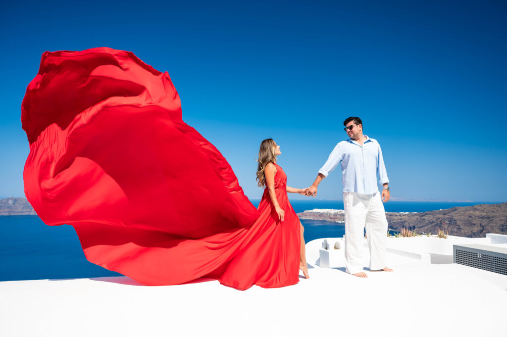 A couple standing on a terrace overlooking the ocean, with the woman wearing a flowing red dress and the man wearing a light-colored shirt and pants. The scenic background features a stunning view of a caldera and the deep blue sea.