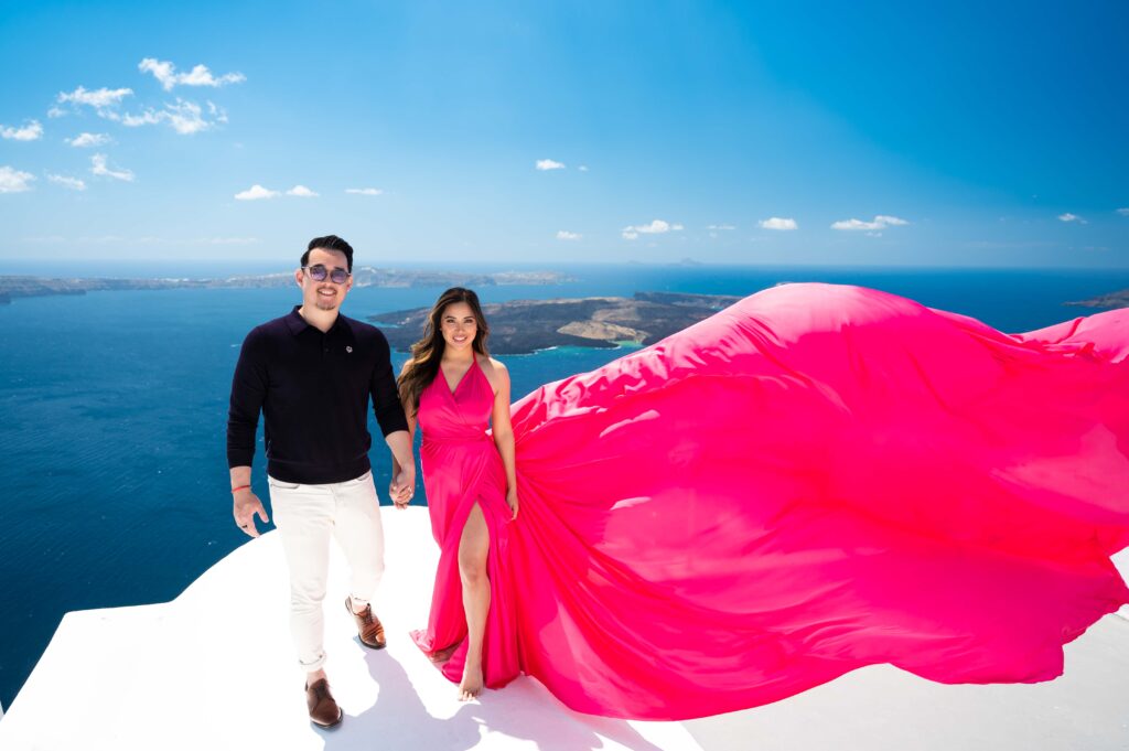 Couple holding hands on a Santorini rooftop, woman wearing a fuchsia flying dress.