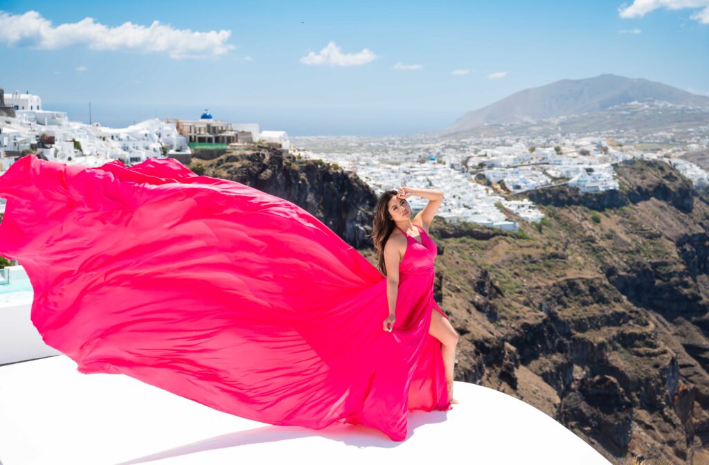 Woman in fuchsia flying dress capturing caldera background while standing on Santorini roof