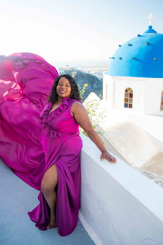 Female smiling to her photographer in purple ruffles attire with Santorini white and blue domes church in the view