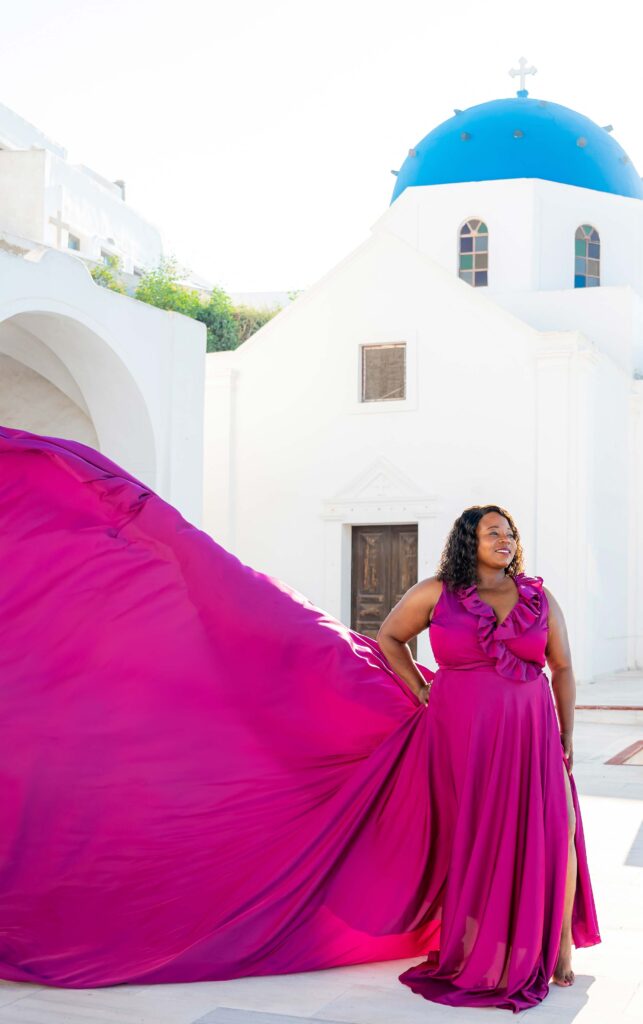 Lady is standing at Blue domes church backyard looking away the sea while wearing a flowy purple dress