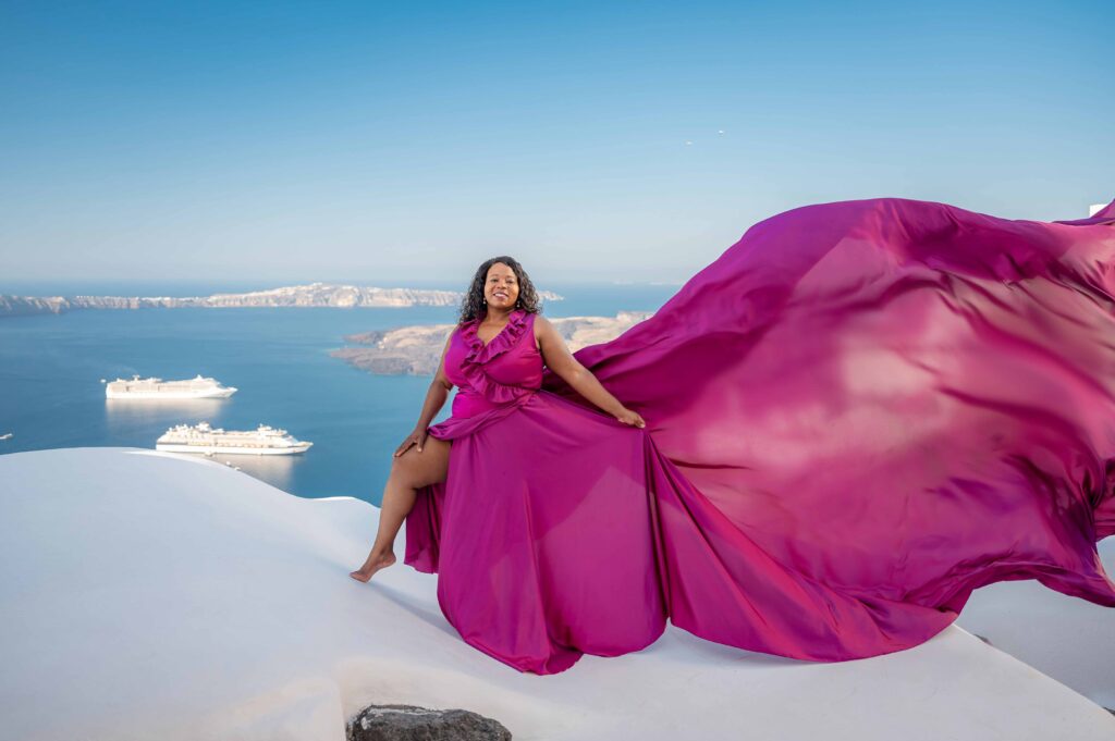 A woman is posing at a white roof wearing purple ruffles dress with a Santorini sea view in the background