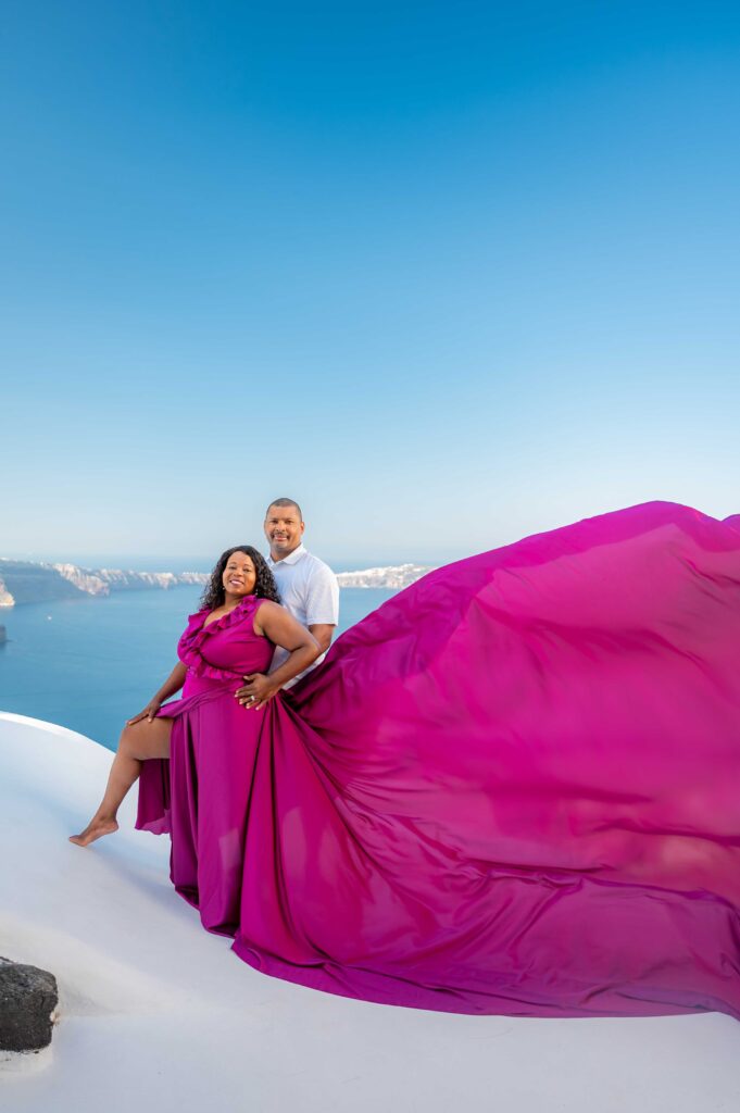 The couple is hugging and looking to the camera, the female wears purple ruffles flowing dress and male white shirt.