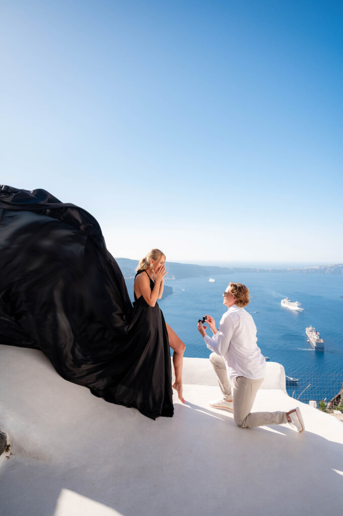 A young gentleman proposing to his girlfriend at Santorini, she looks excited and wears black flying dress