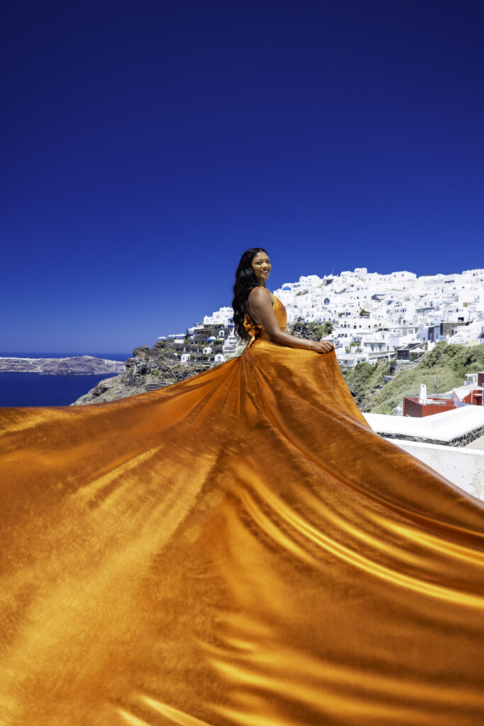 Model twirling in an orange flowing attire on the whitewashed steps of Oia with the Aegean Sea in the background.