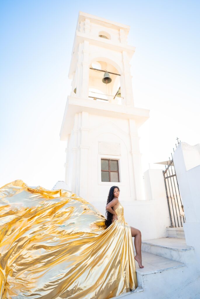 Woman in pose on her hips and in gold flying dress standing with a white tower in the background