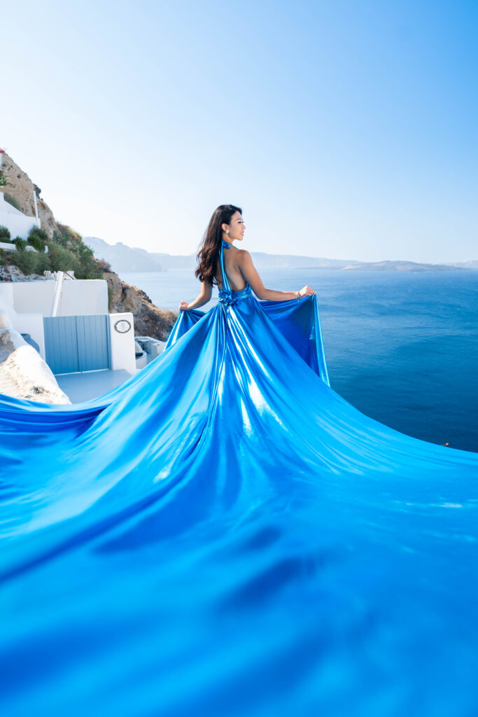Woman in blue metallic flying dress posing on a Santorini cliff with sea view in the background.