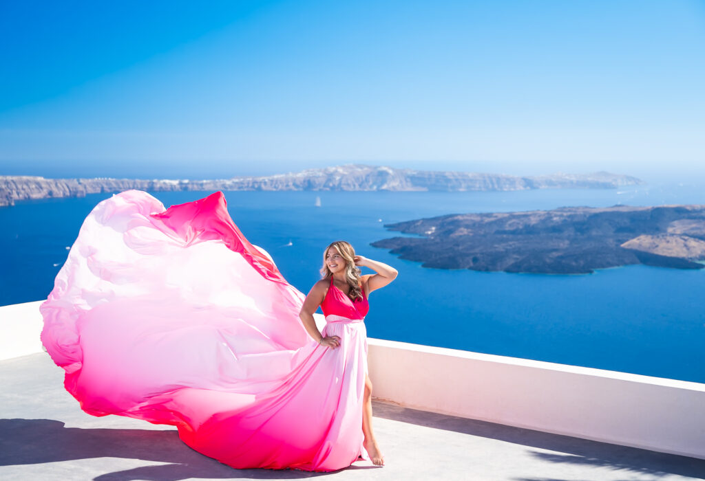 Model in a fuchsia-white ombre flying dress posing on a Santorini rooftop with a high-angle sea view in the background.